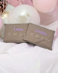 Kirschkernkissen als Wärmekissen, mit beigem Baumwollstoff und goldenen Tüpfchen, personalisiert mit violettem Namen und Geburtsdaten, auf weissem Tuch mit rosa und weissen Kugeln im Hintergrund.