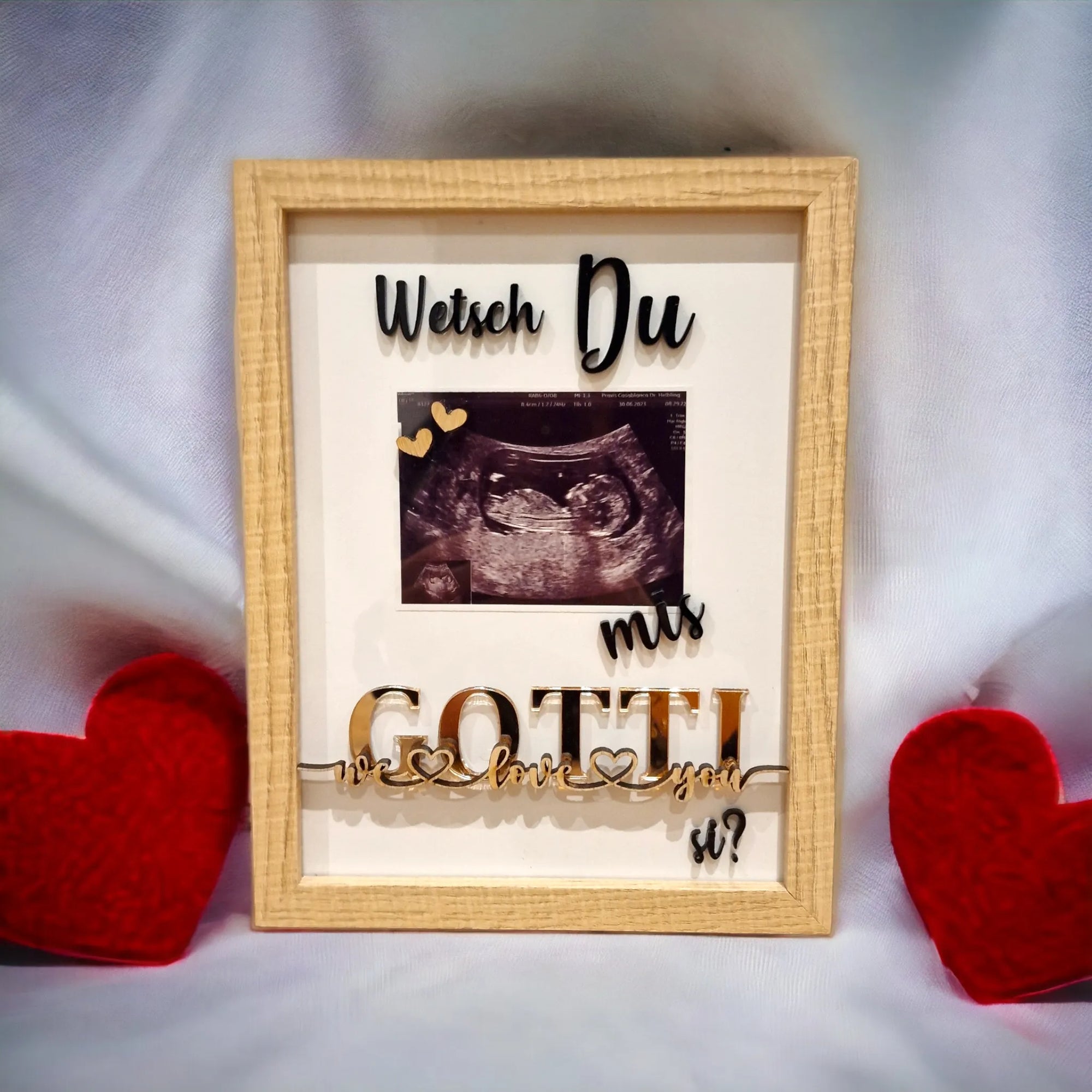Bilderrahmen mit Untraschallbild und Schriftzug “Wetsch du mis Gotti si?“ als Geschenk für zukünftige Gottis, vor weissem Hintergrund mit roten Herzen dekoriert.