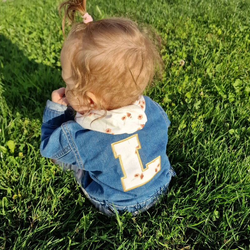 sitzendes-baby-in-wiese-jeansjacke-blau-mit buchstaben-und-dreieckstuch-mit-lina