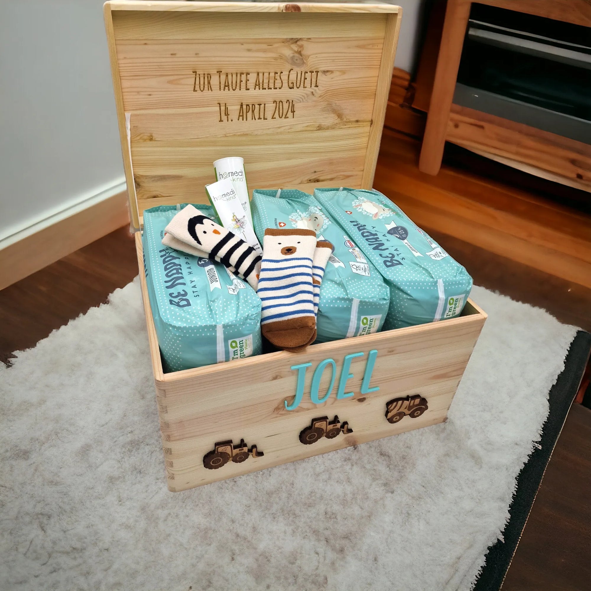 Ein Taufgeschenk für Jungen. Eine Traktot-Motiv Erinnerungskiste, personalisiert mit Namen auf Deckel und Schriftzug auf Innenseite von Deckel. In der Kiste sind Windeln, Socken und Pflegecremen.
