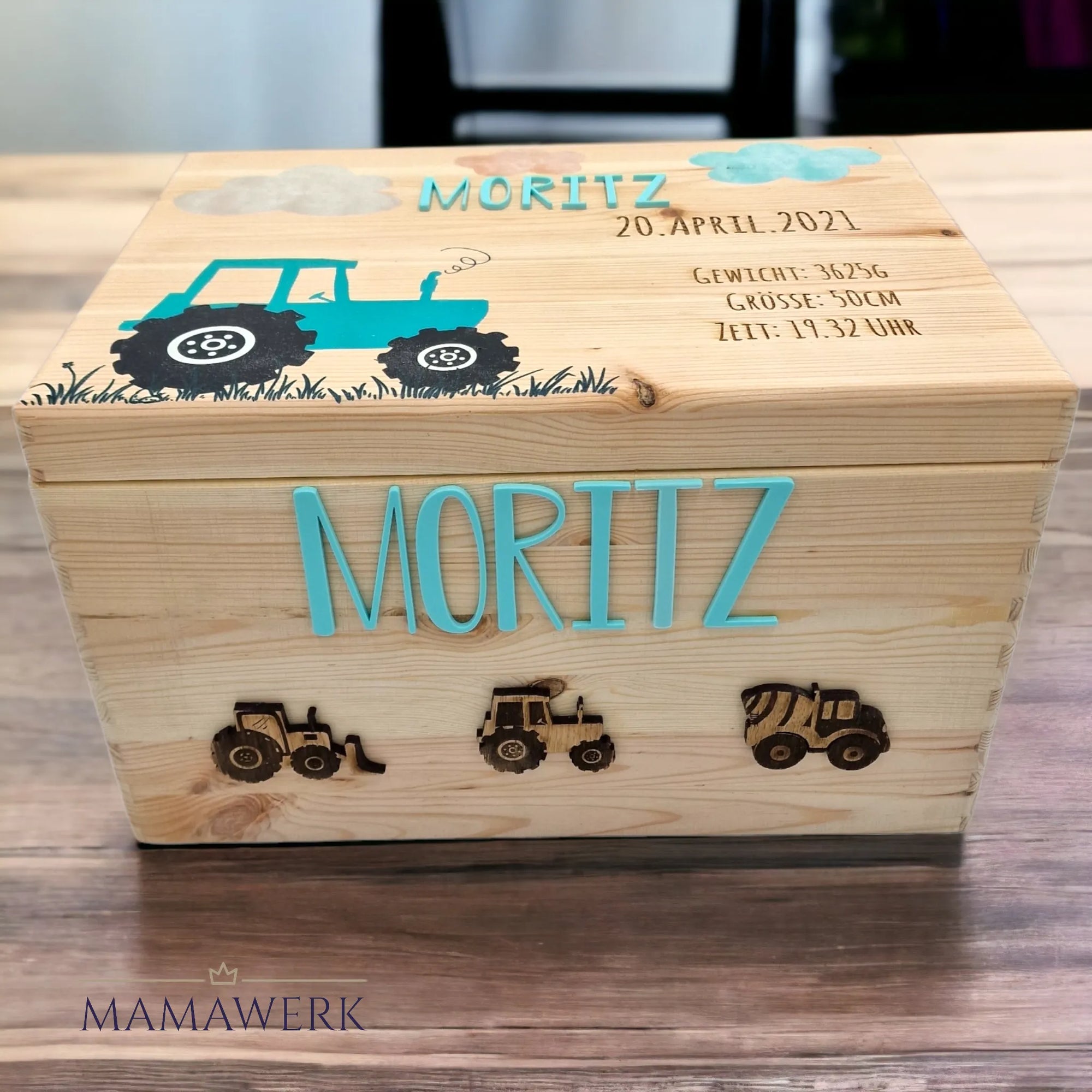 Holzige Erinnerungskiste auf Tisch. Dekoriert mit blauem Traktor auf Deckel, personalisiert mit blauem Namen und Geburtsdaten. An der Seite ebenfalls mit blauem Namen personalisiert und Traktor, Bagger und Betonmischer dekoriert.
