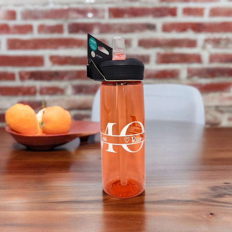 Auf einem Tisch steht eine Orange Camelbak Trinkflasche mit MOM (Mama) Schriftzug und den individuellen Kindernamen mit Herz darüber personalisiert. Schwarzer Verschlussgriff mit durchsichtigem Strohhalm