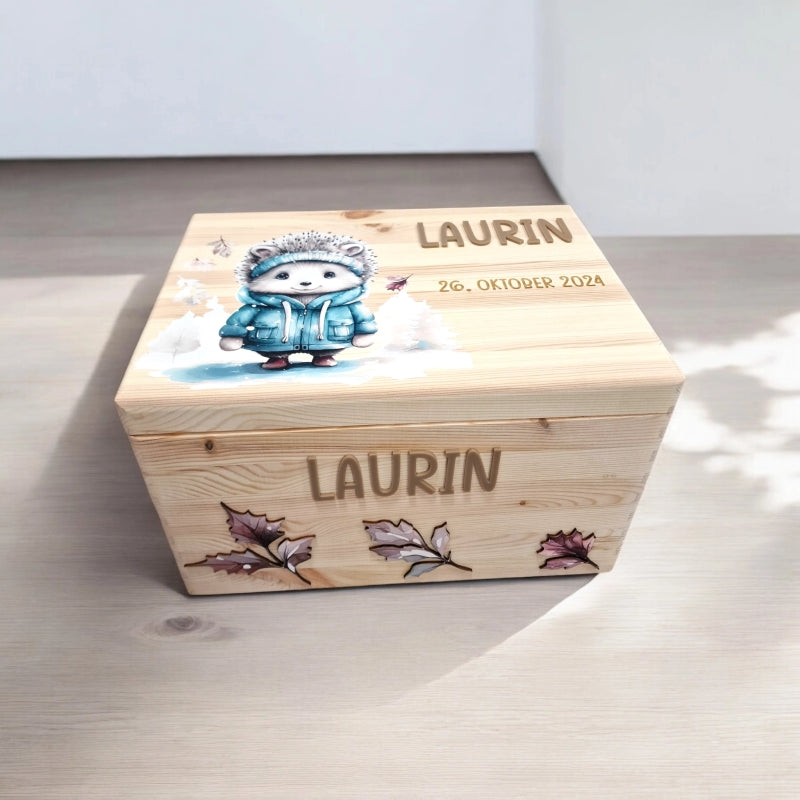 Eine Erinnerungskiste Igel mit Igel-in-Kleidung Motiv auf Deckel gedruckt sowie personalisiert mit farbigem 3D Namen und Geburtsdaten. An der Vorderseite ebenfalls mit farbigem Namen personalisiert sowie mit rosa Blättern dekoriert.