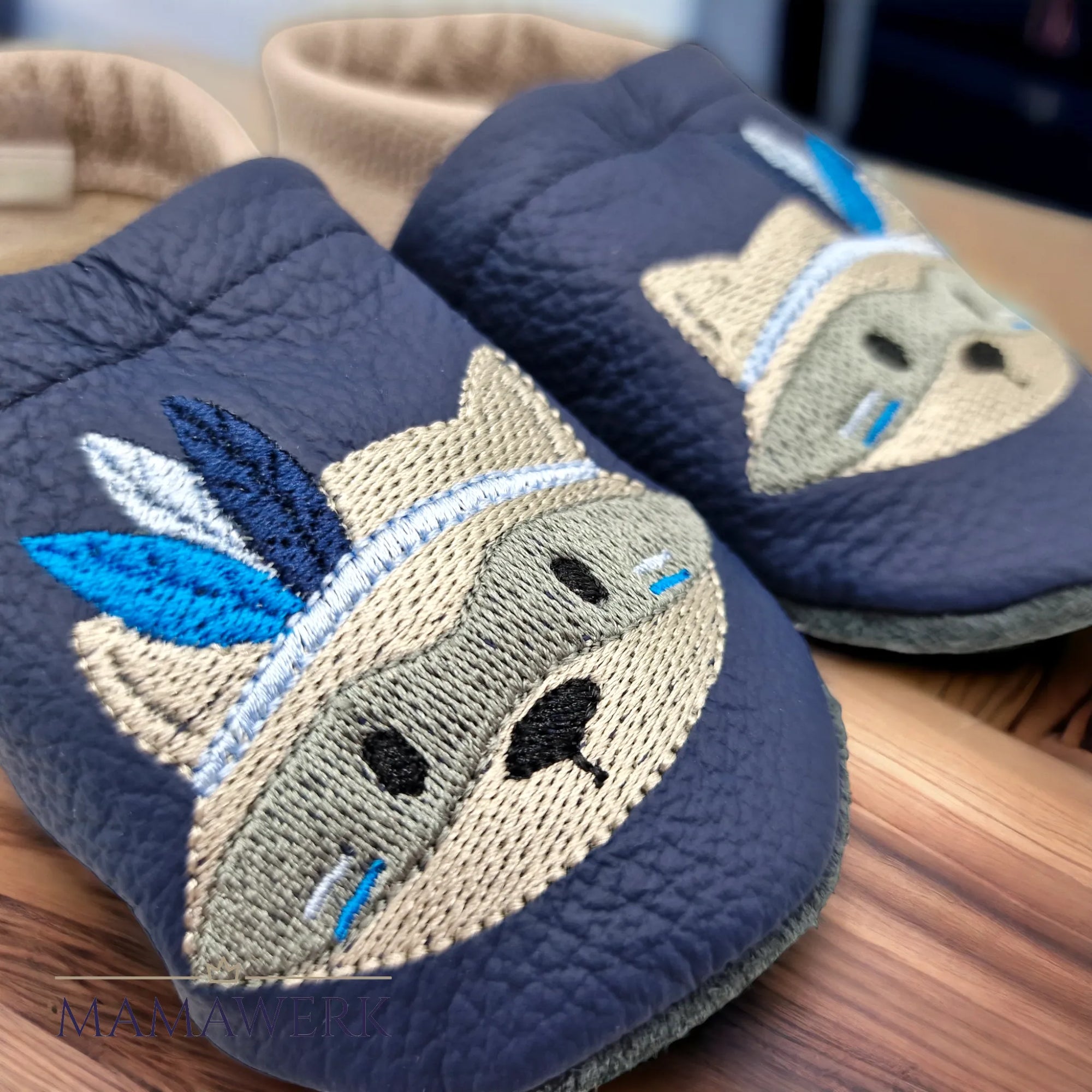 Nahaufnahme von beigen Lederfinken für Kinder mit dunkelblauem Oberleder, welches mit Indianer-Waschbär Motiv bestickt ist. Die Sohle ist aus schwarzem Leder und rutschfest. Die Lederfinkli stehen auf Holztisch.