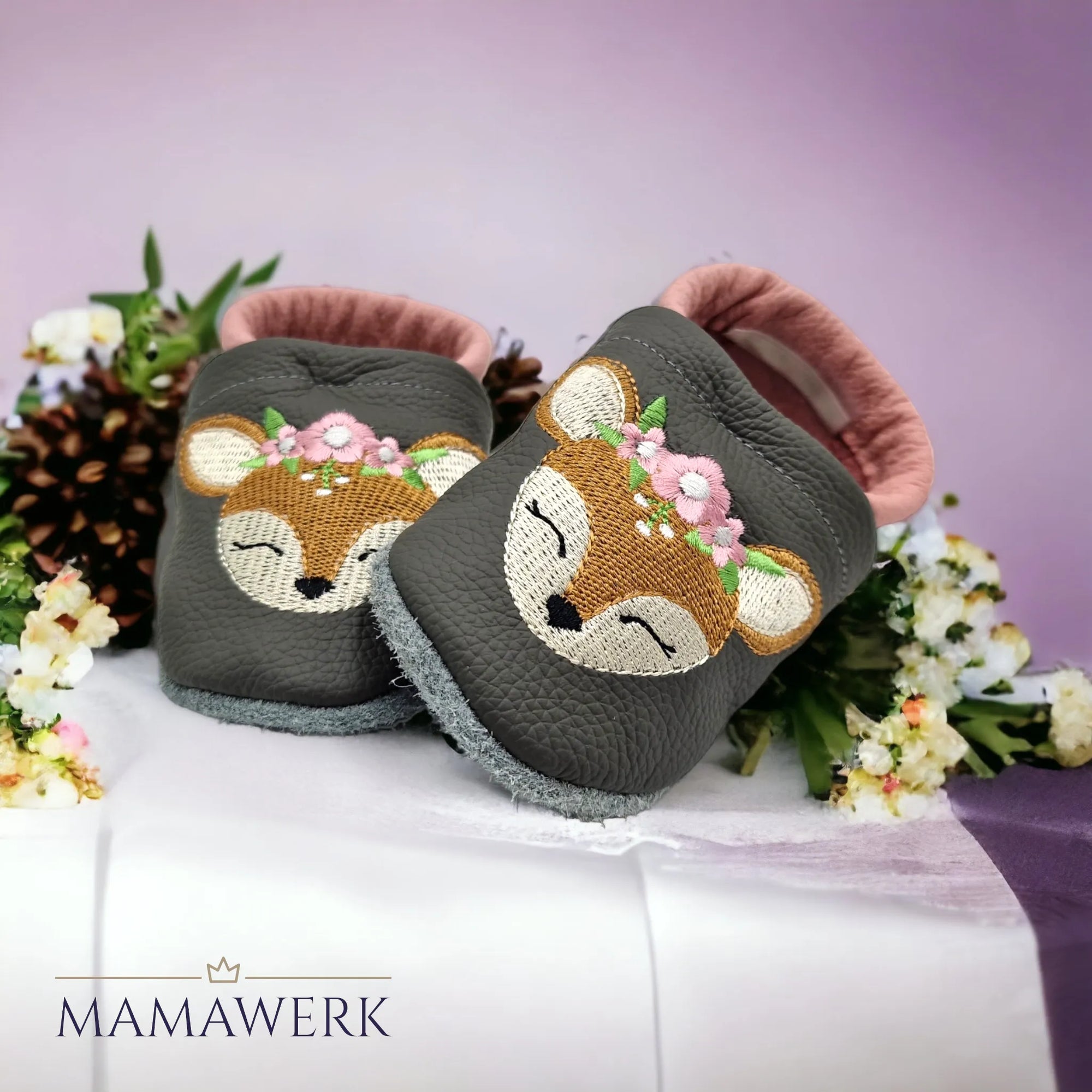 Ein Paar rosa Lederfinken für Kinder mit grauem Oberleder auf weissem Tuch stehend, mit Blumen umrahmt. Krabbelfinken sind mit Boho-Reh Motiv bestickt. Die Sohle ist aus grauem Leder und rutschfest.