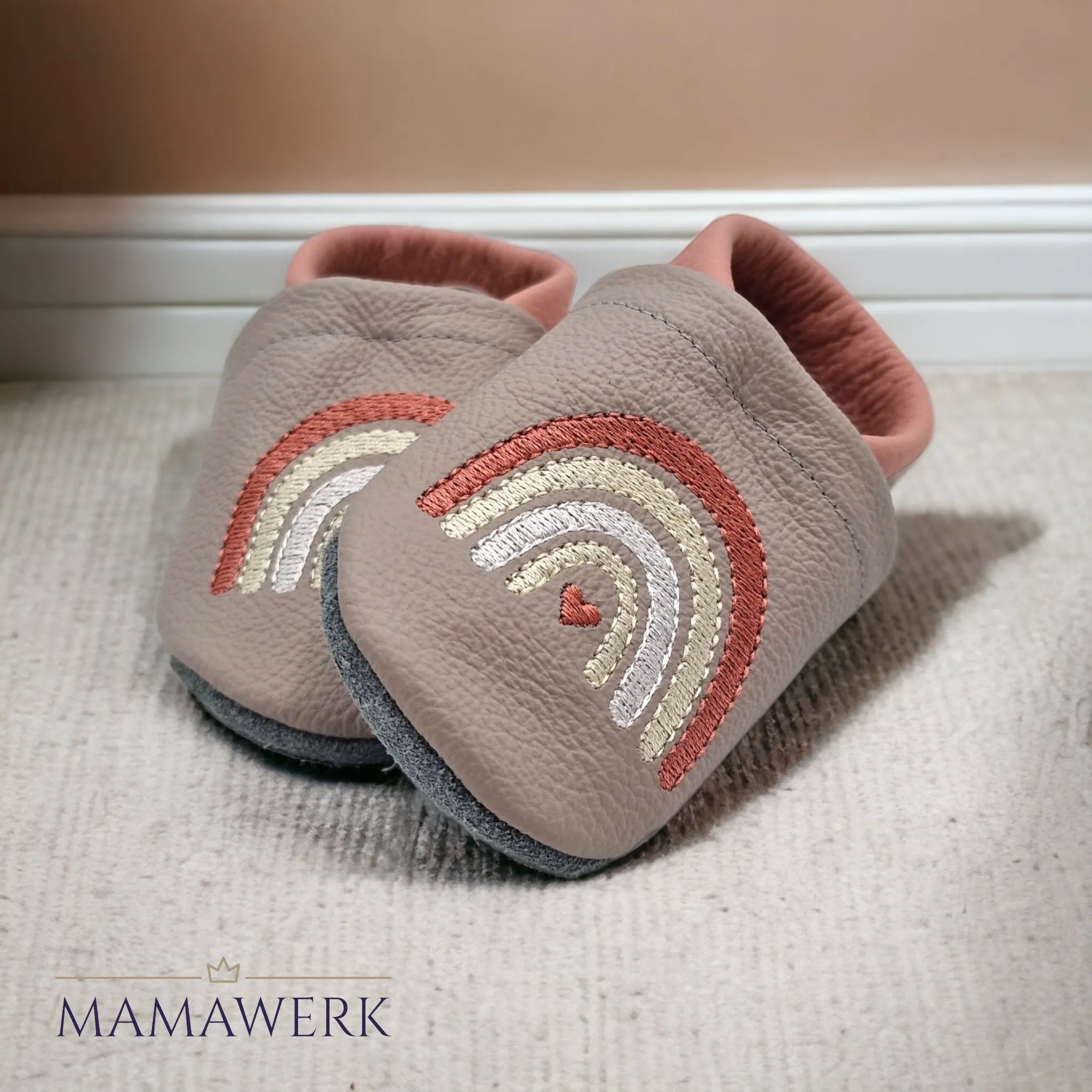 Ein Paar rosa Lederfinken für Kinder mit beigem Oberleder, welches mit Boho-Regenbogen Motiv bestickt ist. Die Sohle ist aus grauem Leder und rutschfest. Die Lederfinkli stehen auf cremigen Teppichboden