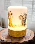 Erleuchtetes LED-Nachtlicht mit Holzsockel und Giraffen- und Elefant-Motiv, mit Namen personalisiert. Auf Holzrondell stehend, vor weissem Vorhang.