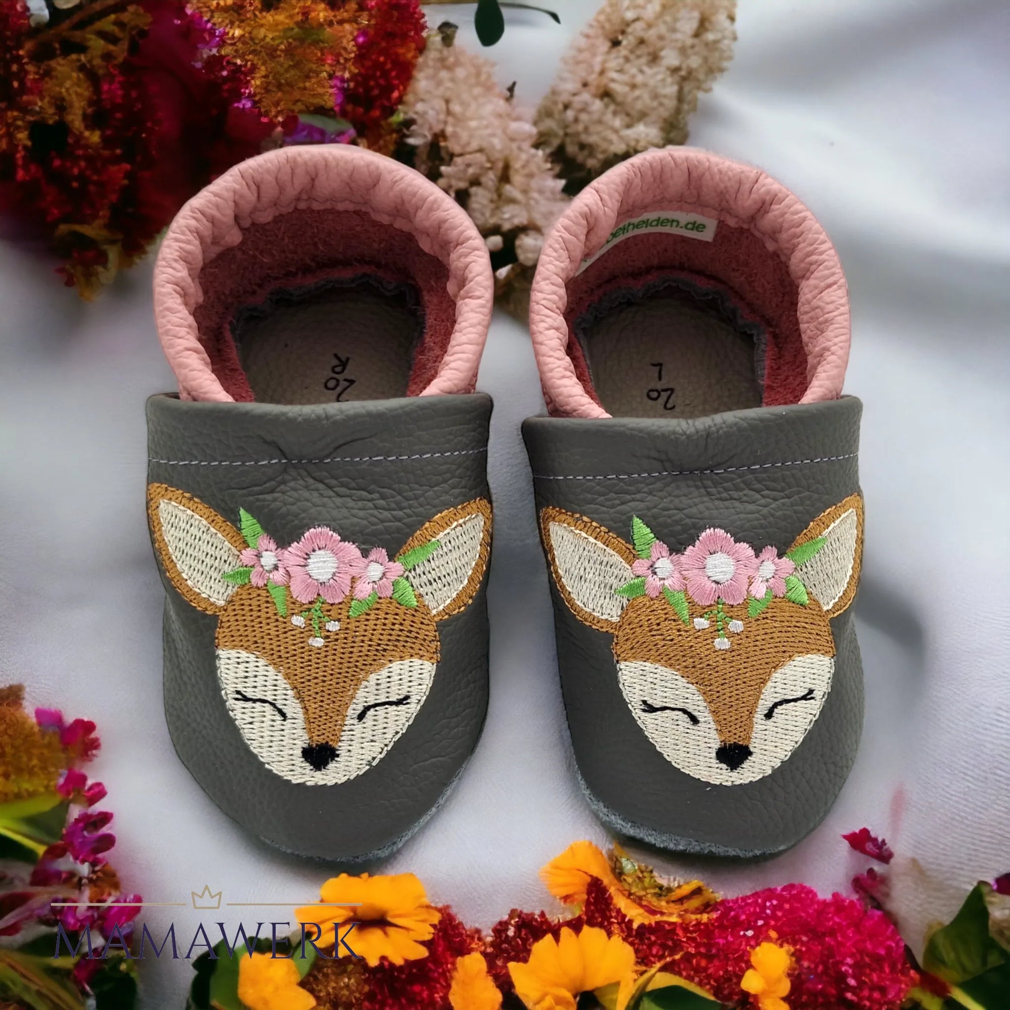 Ein Paar rosa Lederfinken für Kinder mit grauem Oberleder auf weissem Tuch stehend. mit farbigen Blumen umrahmt. Krabbelfinken sind mit Boho-Reh Motiv bestickt. Die Sohle ist aus grauem Leder und rutschfest.