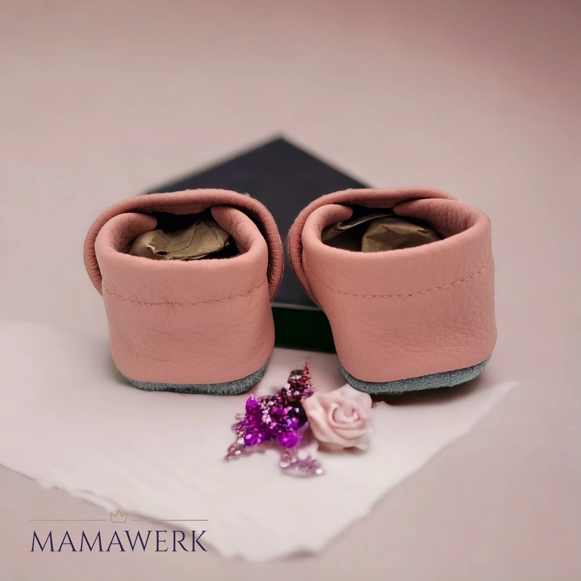 Rückseite von rosa Lederfinken für Kinder mit Spitzbestickung auf Oberleder und Herzbestickung an der Spitze. Die Sohle ist aus grauem Leder und rutschfest. Auf rosa Hintergrund, mit Blume dekoriert.
