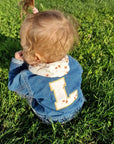sitzendes-baby-in-wiese-jeansjacke-blau-mit buchstaben-und-dreieckstuch-mit-lina