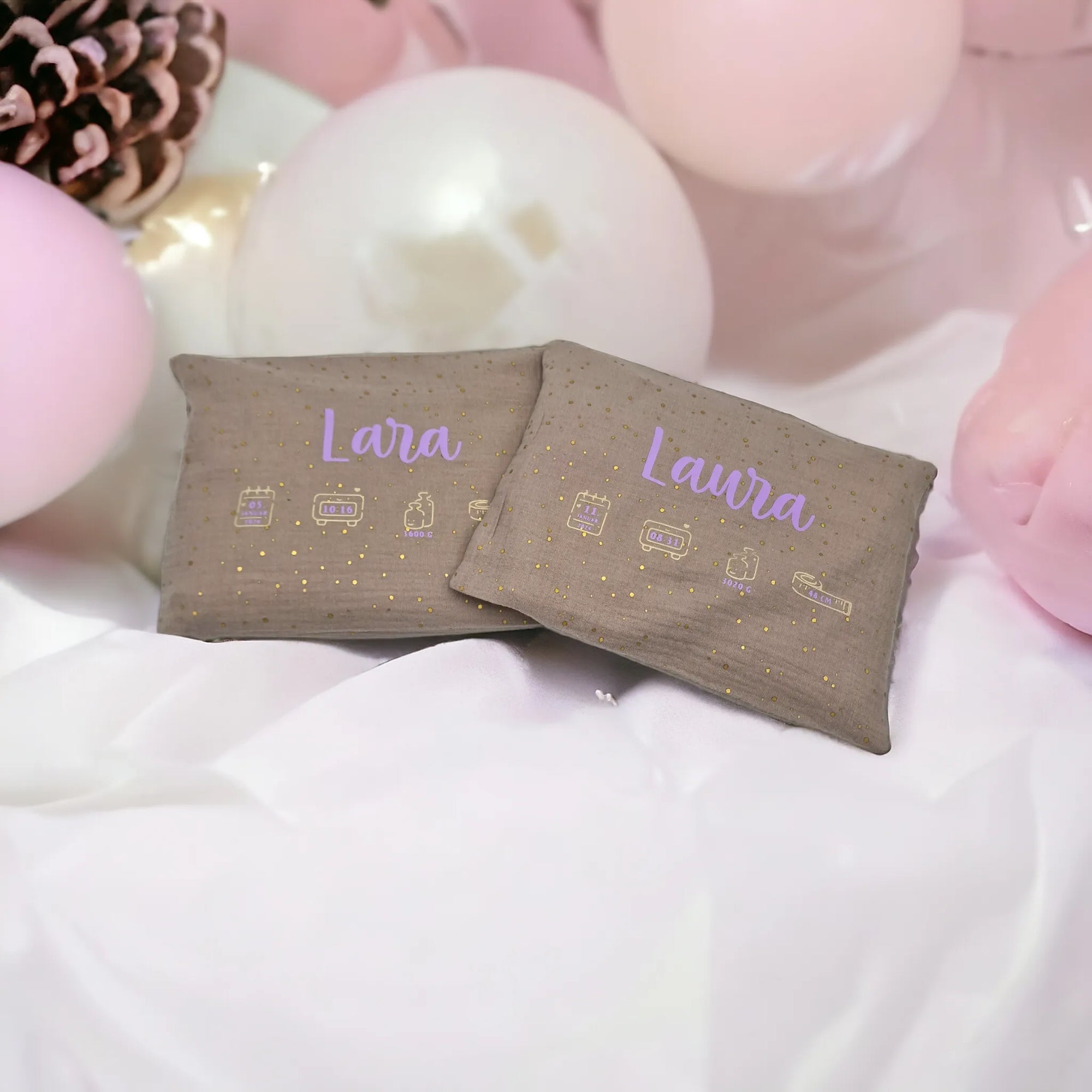 Kirschkernkissen als Wärmekissen, mit beigem Baumwollstoff und goldenen Tüpfchen, personalisiert mit violettem Namen und Geburtsdaten, auf weissem Tuch mit rosa und weissen Kugeln im Hintergrund.